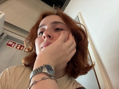 A close-up, upside-down shot depicts a fair-skinned individual with red-brown, wavy hair partially obscuring their face with a hand. The person is wearing a light-colored, possibly beige, top. On their wrist, a silver-toned watch and bracelet are visible. The setting appears to be indoors, possibly a train or bus carriage, suggested by the visible white panels and a safety sign with red pictograms. The image was taken in Lautzenhausen, Germany.