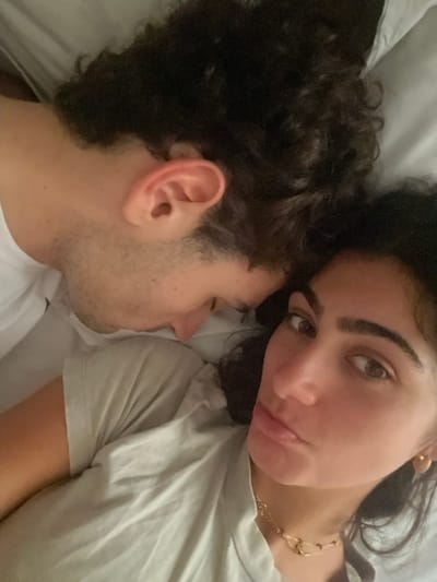 The image captures two individuals lying closely together, likely in a bed with light-colored bedding. On the left, a male individual is depicted in a partial side profile, with his head resting and face turned downwards. He has dark, curly hair, light skin with visible stubble on his jawline, and is wearing a white short-sleeved shirt. His left ear is clearly visible. On the right, a female individual with dark, wavy hair, prominent eyebrows, dark eyes, and full, slightly pursed lips faces the camera directly. She wears a light green/grey short-sleeved top, a layered gold-colored necklace, and a small gold-colored earring in her visible right ear. Her arm, clad in the same light green/grey top, is in the foreground. The ambient lighting appears soft. The image contains no visual information to establish its location in Orléans, France.