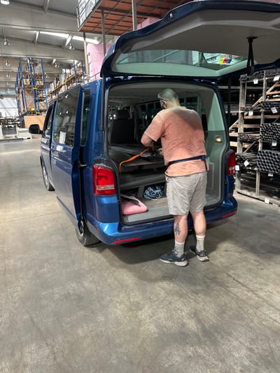 A man with grey hair and wearing a light orange t-shirt, light grey shorts, and grey socks with dark sneakers is standing at the open rear of a dark blue van. He is holding an orange strap and appears to be securing items inside the van. The van's rear hatch and driver-side sliding door are open. The van is parked on a concrete floor within a large warehouse-like building. Racks of metal pipes and other materials are visible on the right side of the image, and shelves stocked with items are in the background on the left. The environment is industrial, with exposed ceiling beams and hanging lights. There is no indication of the specific city of Dub, Montenegro in the image.