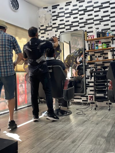 The image depicts the interior of a barbershop in Santa Coloma de Gramenet, Spain. A barber, wearing a black shirt with a white abstract pattern and black pants, is attending to a client seated in a black barber chair. The barber is holding a cape over the client and appears to be in the process of cutting hair. The client is wearing a light-colored mask.
To the left, another individual, possibly a customer or employee, stands wearing a blue and white plaid shirt and denim shorts. They are facing away from the camera, looking towards the barber.
The barbershop features a black and white brick pattern on the wall behind the barber station. Shelves mounted on the wall display various hair care products and tools, including bottles of shampoo, conditioner, and sprays. A large mirror is positioned behind the barber chair, reflecting the interior. A ceiling fan is visible on the wall above.
The floor is covered with grey wood-look tiles. In the foreground, a rolling cart with multiple black drawers holds hairdressing equipment such as a hairdryer and scissors. An electrical outlet with cords plugged in is visible on the wall near the cart.
A display board with images of different hairstyles is mounted on the wall to the left, next to the individual in the plaid shirt. A circular clock with black numbers and hands is mounted high on the wall, showing approximately 10:05. The overall scene suggests a typical, functional barbershop environment.