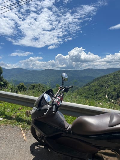 A black scooter is parked on the side of a road overlooking a lush green mountain landscape under a blue sky with scattered white clouds. The scooter is positioned in the foreground, taking up the right half of the image, with its brown seat and handlebars visible. A metal guardrail runs along the edge of the road. The mountainous terrain stretches into the distance, with a winding road visible on one of the slopes. The scene is bathed in sunlight, casting shadows on the road. The image appears to have been taken on a sunny day in Son Dien, Vietnam.