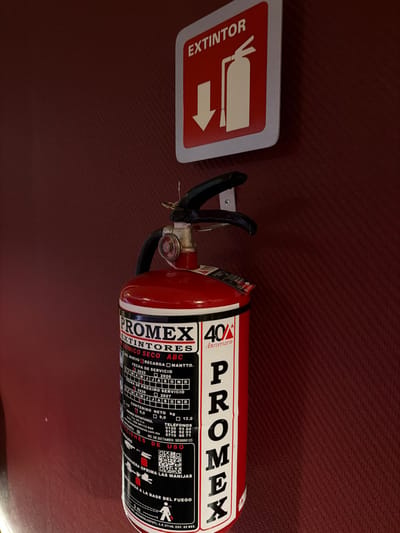 A red PROMEX dry chemical ABC fire extinguisher is mounted on a dark reddish-brown textured wall. Above the extinguisher, a red square sign with a white border displays the word "EXTINTOR" (Extinguisher), a white fire extinguisher icon, and a white downward arrow. The extinguisher's label indicates a service date of January 2023 and a next service date of January 2024. It is a 6.0 kg unit, prominently displaying "40 Aniversario" (40th Anniversary) and contact information including a QR code for a usage manual. The origin or service location listed on the extinguisher's label is Blvd. Peña Flor #456 A, Col. Sauz, C.P. 76117, Querétaro, Qro., México, placing the scene in Santiago de Querétaro, Mexico. No individuals are present in the image.