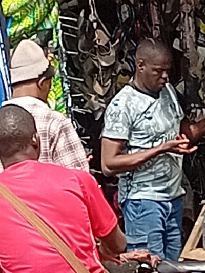 The image depicts an outdoor scene in the city of Ayangba, Nigeria, at what appears to be a vendor stall. In the foreground, positioned with their back to the viewer, is an individual wearing a red short-sleeved shirt with a brown strap diagonally across their back. Directly behind them, slightly to the left, and also facing away from the viewer, is another individual wearing a light-colored knit cap and a light-colored, possibly plaid, long-sleeved shirt. To the right, a third individual with dark skin and short hair stands, facing downwards and slightly to the left. This person is wearing a light grey patterned short-sleeved t-shirt and blue denim shorts or jeans, with their hands held up in front of them, palms inward, as if examining or manipulating small items. The background features a dense vertical display of dark-colored merchandise, possibly footwear or small bags, hanging from a structure. To the far left in the background, a green and yellow patterned fabric is visible. Partially obscured dark objects are present on a surface in the lower right foreground, adjacent to the individual in the grey shirt. No direct interaction between the visible individuals is clearly depicted.