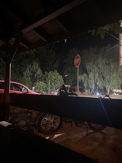 In Lozenitsa, Bulgaria, at night, a scene unfolds under a wooden canopy. In the foreground, the rear wheel of a dirt bike is visible, its spokes catching the dim light. Behind it, a section of a red car is parked. Further back, a red octagonal stop sign stands beside a large, leafy tree. The background is filled with various trees and vegetation, creating a somewhat wild and overgrown ambiance. A person, partially obscured, appears to be sitting or leaning near some objects, including what looks like another bicycle. The lighting is low, suggesting it is late evening or early morning.