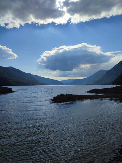 This is a photograph of a wide, calm lake surrounded by lush green, tree-covered mountains. The sky is a vibrant blue, with scattered white and grey clouds. Sunlight streams through a gap in the clouds, creating visible rays of light. The water has a rippled surface, reflecting the sky and the surrounding landscape. There are small, dark landmasses with sparse vegetation along the edges of the lake, and the shoreline appears rocky. The scene suggests a natural, serene environment, possibly a remote natural area. While the prompt mentions Kordon Baigazan, Russia, this specific geographical context cannot be determined solely from the visual information within the image itself.