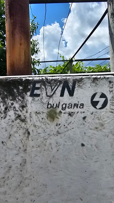 The image shows a weathered, white, textured surface with "EVN bulgaria" written in dark lettering. To the right of the text is a circular logo with a lightning bolt symbol. The surface appears to be part of an outdoor utility box or structure. In the background, there is a rusty pipe on the left, green foliage, and a bright blue sky with white clouds. The overall scene suggests an outdoor urban or semi-urban environment, possibly in Karnare, Bulgaria, given the inscription.