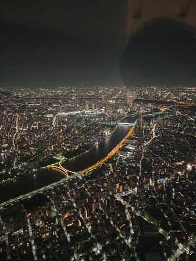 An aerial view captures the cityscape of Sumida, Japan, at night. The Sumida River cuts through the city, with multiple bridges illuminated with orange lights spanning across it. The buildings are dense, with scattered lights resembling stars. A dark shape, possibly a window reflection, appears on the upper left side. The image is slightly rotated counter-clockwise.