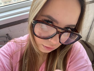 A person, oriented upside down, is wearing eyeglasses with tortoise-shell frames and a silver nose ring. They are wearing a light pink top. The subject's hair is blonde with darker roots. Visible behind the person is a window and the exterior of a building, suggesting an interior setting. The location is Prigorodny, Russia.