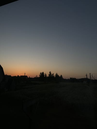 The image captures a rural scene at dusk in Chemerne, Ukraine. The sky transitions from a deep blue at the top to a warm orange hue near the horizon, indicating the time of day is sunset or twilight. Silhouetted against the fading light are trees, power lines with utility poles, and the rooftops of several buildings. The foreground is dominated by a dark, densely vegetated area, possibly a field or garden, with the outlines of structures suggesting a residential or agricultural settlement. The overall mood is tranquil and serene, characteristic of the end of a day in the countryside. There is no visible text in the image.