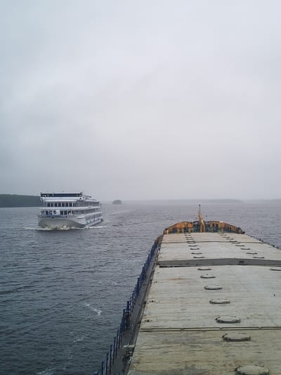 A photograph shows two ships on a body of water under a cloudy sky. The first ship, a passenger liner, is white with multiple decks and is moving towards the viewer, leaving a wake. In the foreground is the deck of a barge, gray and weathered, with evenly spaced round hatches. In the distance, there is a landmass with trees. The water is choppy and gray. According to the metadata the location of the photo is in Topornya, Russia.
