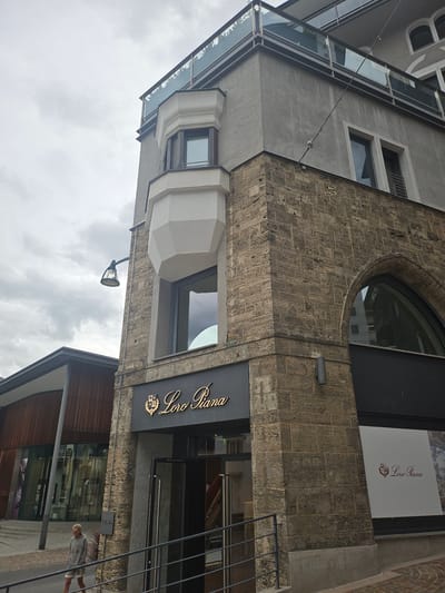 This is a vertical, medium shot of a building's facade in St. Moritz, Switzerland. The building features a mix of architectural styles. The upper portion has a modern, glass-enclosed section that angles upwards, contrasting with the rough, textured stone facade below. A prominent circular window is set into the stone. A sign with the text "Loro Piana" in gold lettering is mounted on a dark surface above a shop entrance. A large poster or advertisement featuring "Loro Piana" with a logo is visible in an upper window. The scene appears to be outdoors, with a cloudy sky suggesting overcast weather. A street lamp is visible attached to the building. In the lower right of the frame, a person is walking on a paved area in front of the shops. The overall impression is a street view of a commercial building in an urban or semi-urban setting.