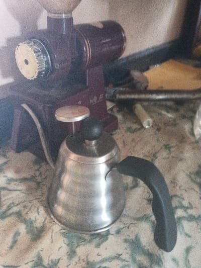 The scene, set in Dogsenang Gorphoog, Bhutan, depicts a close-up view of coffee preparation equipment positioned on a mottled green and off-white patterned surface, likely a countertop or table. In the foreground, a brushed stainless steel gooseneck pour-over kettle with a black ergonomic handle and a black lid knob rests on the patterned surface. Directly behind and slightly to the left of the kettle is a maroon-colored electric burr coffee grinder, featuring a translucent bean hopper on its top and a prominent silver adjustment dial on its left side. Faint Chinese characters are discernible on the grinder's lower front. The background to the right of the equipment appears cluttered, with a visible metal bar or pipe and a small, light-colored cylindrical object resting on a beige or tan surface. No individuals are present.