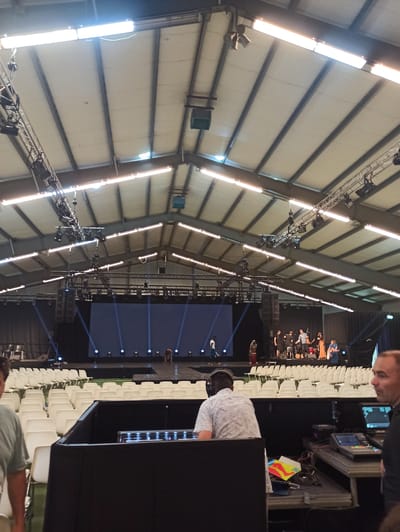 The image shows a wide shot of a large indoor venue, likely an auditorium or conference hall, with a high, arched ceiling supported by metal beams and covered with insulated panels. Long fluorescent lights run along the ceiling's structure, illuminating the space. In the foreground, a person wearing headphones is seated and looking down at a console with a screen, suggesting they are operating technical equipment for an event. To the left and behind this person are rows of empty white chairs, indicating a seating area for an audience.
Further into the venue, a stage area is visible with a large dark screen displaying blue light beams projected onto it. A group of people are on the stage, some appearing to be performers or speakers, with musical instruments and equipment present. Another person, seen from the side, is standing near the stage. The overall scene suggests an event is about to begin or has just concluded, given the presence of both technical staff and performers, and the empty seating. The lighting appears to be artificial, typical of an indoor event setting. No specific time of day or weather conditions can be inferred from the image. The location is identified as Warffum, Netherlands.