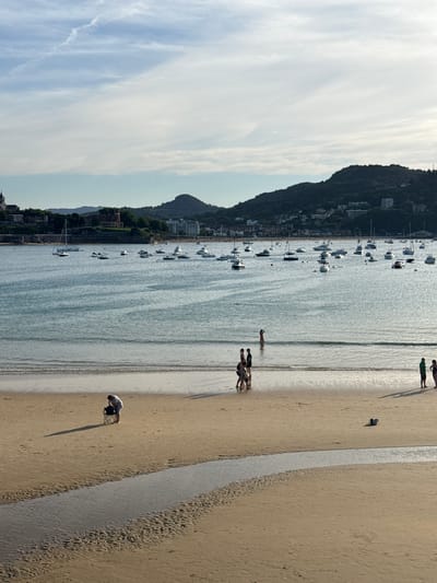 The image depicts a scenic view of La Concha Bay in San Sebastián, Spain, at what appears to be late afternoon. The foreground is a sandy beach with a small stream of water flowing across it. In the mid-ground, several people are visible on the beach. To the left, a person in a wheelchair is looking down at something on the sand. Further to the right, a group of people are walking along the shoreline. In the background, the calm waters of the bay are dotted with numerous boats of various sizes, including sailboats and motorboats. Across the bay, the lush green hills of San Sebastián rise, with buildings and a prominent church spire visible on the slopes. The sky is partly cloudy with soft sunlight casting shadows on the beach.