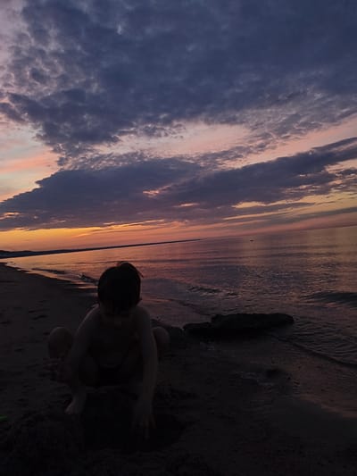 At dusk on a beach in Apšuciems, Latvia, a young child, seen from behind and in silhouette, crouches in the sand. The child is partially obscured by shadow, with their arms bent and hands near a pile of sand. The beach is dark and sandy, with the gentle waves of the Baltic Sea lapping at the shore. In the distance, the sea stretches to the horizon under a sky painted with the soft hues of sunset, transitioning from warm oranges and pinks near the horizon to deeper blues and purples as they ascend. Wispy clouds are scattered across the sky, their undersides catching the fading light. A small, dark object, possibly a boat or buoy, is visible as a tiny speck on the calm water far from shore. To the right of the child, a dark, elongated object lies partially submerged in the shallow water at the edge of the sand.