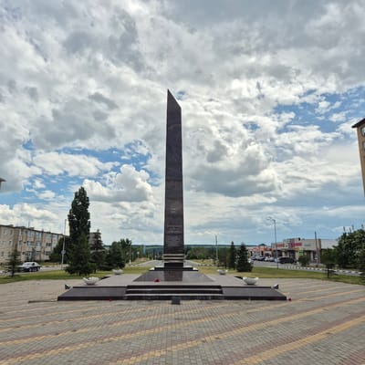 This is a frontal, eye-level, wide-angle photograph of the obelisk monument located in the city of Liski, Russia. The monument is a tall, slender, dark obelisk with a sharply angled top, positioned centrally in the frame. It stands on a multi-tiered, dark stone base with steps leading up to it. Two white, boat-shaped planters are visible on either side of the base, and a single red rose lies on the lower steps of the monument.

The monument is situated in a paved plaza with a distinctive pattern of reddish-brown and yellow rectangular paving stones. In the background, to the left, is a multi-story apartment building constructed of light-colored brick. To the right of the monument, a street is visible, lined with trees and shops. One shop has a sign that reads "КОСМЕТИК". Cars are parked along the street and a dark sedan is driving on the road. The sky is filled with dramatic, fluffy white and grey clouds against a backdrop of blue sky. Several tall, dark green evergreen trees are planted around the plaza and in the distance. The overall scene depicts a public space, likely a memorial or park, in a modern urban or suburban setting.