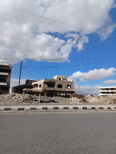The image displays an urban scene in Al-Qadam Municipality, Syria, depicting widespread structural damage under a partly cloudy blue sky. In the foreground, an empty asphalt road extends horizontally, demarcated by a curb composed of alternating dark and light gray rectangular blocks. Beyond the curb, a substantial accumulation of concrete and building rubble forms an uneven ground surface, with sparse, dry vegetation visible in some areas.

Three multi-story concrete structures are prominent. On the left, a partially visible building shows significant damage with missing wall sections and exposed interior elements. Central to the frame is a severely damaged, multi-story concrete building exhibiting extensive structural collapse, numerous impact perforations on its remaining facades, and exposed rebar. Its lower sections are largely obscured by a large mound of debris. Behind this central structure, a black utility pole rises, with multiple dark utility lines traversing the upper portion of the sky, some appearing as dashed segments. To the far right, another concrete building, appearing either unfinished or significantly damaged, is partially visible. No individuals are present, and no actions or interactions are depicted within the scene. The background reveals additional urban structures along the horizon line, consistent with a city environment.