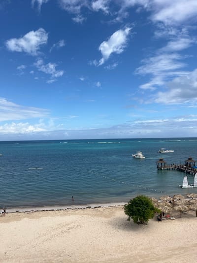 A vibrant coastal scene shows a blue sky dotted with fluffy clouds. Turquoise water stretches to the horizon, marked by a pier and boats. A white sand beach features palm umbrellas and a solitary tree. A few people are visible on the shoreline.