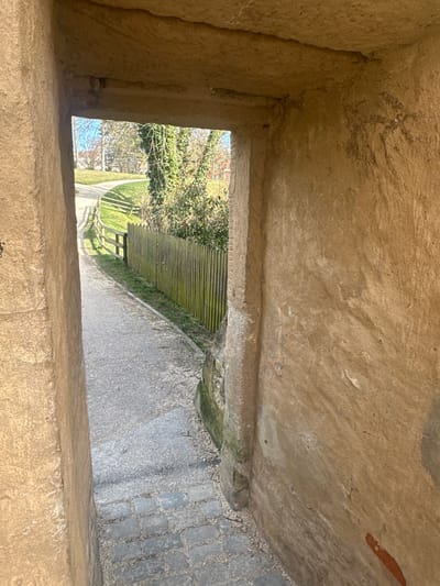 The image displays a view from inside a stone archway or passage looking outwards onto a curving path and a landscaped area, located in Weißenburg in Bayern, Germany. The archway's interior walls are constructed from rough, light brown or beige stone masonry, showing weathering and a textured surface. The ground within the archway transitions from grey cobblestones at the immediate foreground to a light grey gravel or compacted dirt path leading away.
Beyond the archway, the path curves gently to the left and is bordered on the right by a weathered wooden picket fence, partially covered in green moss or algae. Adjacent to the fence are grassy areas and various trees and shrubs, including some with vibrant green foliage and others appearing deciduous with bare branches against a clear blue sky. A low wooden rail fence runs along the left side of the path further into the scene. The exterior is brightly lit by sunlight, casting subtle shadows, while the interior of the archway is in comparative shade. No people, animals, or active subjects are depicted.