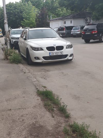 The image displays an outdoor scene in Lom, Bulgaria, featuring several parked vehicles and a building under a tree canopy during daylight. In the foreground, a white BMW 5 Series Touring (E61) is parked on an unpaved shoulder of dirt and gravel, facing the viewer. Its front license plate displays 'CB 1021 TK', and minor damage is visible on the lower right side of its front bumper. Immediately behind the BMW, a white Dacia Duster is partially visible, parked on the same shoulder next to a metal utility pole covered with various faded papers.
Beyond these vehicles, in a separate unpaved parking area, several other cars are parked. These include, from left to right: a grey Renault Scenic (first generation) with the license plate 'M 0089 BH', a silver Mitsubishi Outlander (first generation) displaying license plate 'M 2902 BP', and a black SsangYong Rexton with the license plate 'CA 4259 MM'. Behind these parked vehicles stands a white, single-story building featuring a dark-colored roof, two small rectangular windows, and an exterior air conditioning unit mounted on its wall.
The ground surface in the immediate foreground consists of uneven dirt, gravel, and fragments of concrete, with small patches of green vegetation and weeds growing along a broken concrete curb. Large green trees form a dense background, obscuring the horizon. No people are present in the scene.
