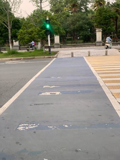 The image shows a crosswalk with a green light on the traffic signal. There is a person riding a scooter on the crosswalk. The crosswalk is a marked pedestrian crossing, with the surface painted blue and yellow, and is located at the intersection of a street and a pedestrian path. The crosswalk has some cracks and wear and tear. The scene is located in Wuhan, China, near the coordinates 30.51°N 114.404°E. There are trees and plants in the background, with concrete sidewalks and a white curb.