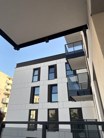 The media file shows an upward-facing view of modern architecture in Białystok, Poland. The scene features a building with a white facade accented by black frames around the windows. Several windows are visible, some reflecting the sky, others showing interior spaces, and one reflecting trees. There are also balconies with glass railings. To the left of the frame, a textured, light-colored surface is angled upwards, suggesting an overhang or the underside of a structure. In the background, the sky is clear and blue, indicating good weather and likely daytime. The overall impression is one of contemporary urban design. There is no visible text in the image.