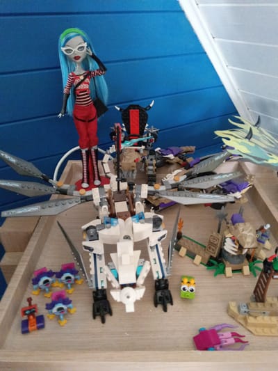This image displays a collection of toys on a wooden surface, likely a shelf or table. In the background, a blue, textured wall is visible, possibly made of wood panels. The overall setting appears to be an indoor play area or display.

The primary subjects are various toys, including a fashion doll with blue hair, white glasses, and a red and black striped outfit. Next to the doll is a complex, multi-colored LEGO build, resembling a mechanical dragon or creature with wings and horns. In the foreground, there are smaller LEGO constructions, including several bug-like figures with purple and pink accents, a red and orange robot-like figure, a small green character with large googly eyes, and a pink and purple octopus-like toy. Several other LEGO pieces, some resembling desert structures and a ladder, are scattered around the larger builds.

The toys are arranged in a static display, suggesting no particular activity is taking place. The lighting appears to be artificial and somewhat diffused, indicating it might be daytime indoors or evening. There are no visible signs of weather conditions or specific time of day cues beyond the general indoor lighting. No text is visible in the image. The composition and arrangement of the toys imply a personal collection or a play setup. The location context of Lesnyye Polyany, Russia, is provided but not visually inferable from the image itself.