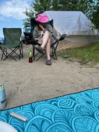 A person wearing a pink cowboy hat sits in a camping chair on a sandy area, looking at a phone. A bottle of Lipton iced tea is on the ground next to them. In the background, a large white tent is set up, and trees are visible. A green camping chair is also present. A can and a silver object are on a blue patterned mat in the foreground. The scene appears to be a campsite.