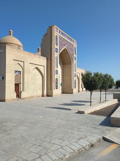 The image depicts a view of the courtyard of the mausoleum of Khwaja Abu Nasr Parsa in Kasri Arifan, Uzbekistan. The mausoleum, constructed from light-colored bricks, features a prominent dome and an ornate arched entrance adorned with blue and white tilework. In front of the mausoleum, a paved courtyard is visible, with several young trees planted in a raised bed along the right side. The sky is a clear, vibrant blue. The scene is empty of people, save for a single motorcycle in the distance. The overall impression is one of serene architectural beauty in a sun-drenched environment.
