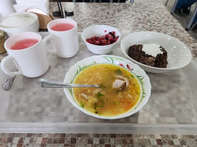 A tray of food sits on a checkered table. On the tray are two white mugs filled with a pink beverage, a white bowl containing a beet salad, a white bowl with a portion of what appears to be meatloaf topped with sour cream, and a large bowl of chicken noodle soup with visible pieces of chicken, noodles, and vegetables. A spoon rests in the soup. In the background, a sugar dispenser and salt and pepper shakers are visible. The scene appears to be indoors, possibly in a cafeteria or dining hall, with a hint of a patterned tablecloth or placemat on the table. The lighting suggests it could be daytime. No people are directly visible, but parts of other diners' legs and clothing are visible in the background, indicating a public eating space. The overall impression is a simple, practical meal.