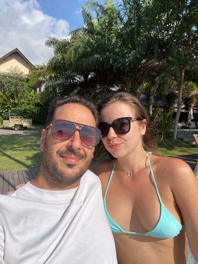 In Buleleng, Indonesia, a man and a woman are captured in a close-up selfie outdoors. The man, on the left, wears a white t-shirt and large, dark-rimmed sunglasses, with a beard and short dark hair. The woman, to the right, sports a light blue bikini top and dark sunglasses, with her light brown hair falling over her shoulders. She wears a delicate silver necklace with a circular pendant. The background reveals lush green grass, palm trees, and the edge of a building with a brown roof. A small utility vehicle is visible on the grass to the left, and in the distance, lounge chairs and umbrellas suggest a poolside or resort area. The sky is blue with scattered white clouds.