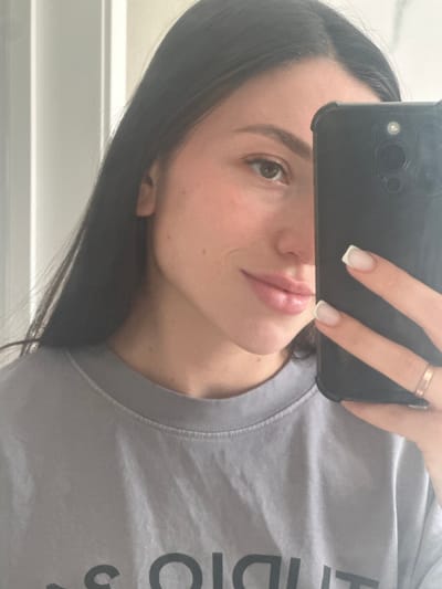 In an indoor setting, a young woman with long, dark hair takes a selfie with her phone. She is wearing a grey t-shirt. The phone, a black iPhone, is held in her right hand, obscuring the right side of her face. Visible on her hand are clean, French-tipped fingernails and a rose gold ring on her right ring finger. Her left eye, eyebrow, and part of her nose and lips are visible. The background appears to be a light-colored wall with vertical lines, possibly indicating a door or partition. The lighting is soft and appears to be natural or diffused artificial light. There is no indication of the specific location within Эстачи, Russia.