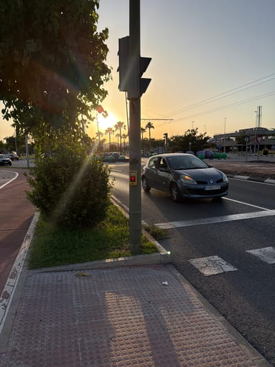 This image captures an outdoor scene in Castelló de la Plana, Spain, during what appears to be late afternoon or early evening. The setting is a street with a pedestrian crossing and a sidewalk. A dark gray Renault Clio is stopped at the pedestrian crossing, suggesting traffic or a waiting period.

The sun is low in the sky, casting a warm, golden light and creating lens flare effects, particularly through the foliage of a large tree in the foreground. The sky is a soft gradient of blue and orange, indicative of sunset. Palm trees are visible in the background, a common sight in this region. The street is paved with asphalt, and the pedestrian crossing is marked with white lines. The sidewalk is made of reddish-brown tiles with a circular pattern.

A traffic light pole is prominent on the right side of the image, with a pedestrian crossing button visible on it. The button is illuminated red, indicating "don't walk." In the distance, construction cranes and other structures suggest urban development.

There is some visible text on the license plate of the car: "0985 FFK". A small, red sign on the traffic light pole also displays text and symbols, likely related to pedestrian crossing instructions. The overall mood is peaceful and serene, bathed in the warm glow of the setting sun. The presence of trees and grass adds a touch of nature to the urban landscape.