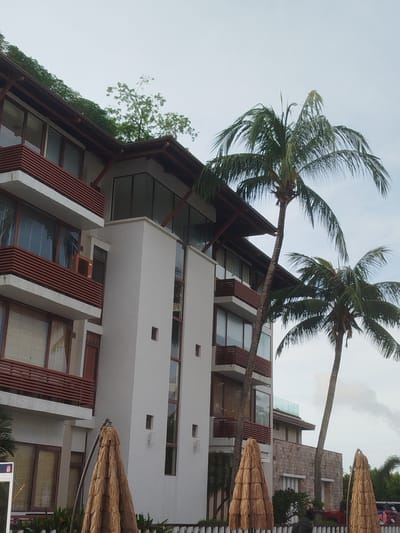 The image shows a modern, multi-story building with a white facade and dark wood trim. The building features multiple balconies with horizontal slatted railings and large windows. Several palm trees are visible in the foreground and middle ground, with their fronds reaching towards the sky. Two straw-covered umbrellas are set up in front of the building. In the background, another building with a stone facade can be seen. The sky is overcast. The scene appears to be a resort or hotel in a tropical location, likely Ko Kaeo, Thailand, given the presence of palm trees and tropical architecture. There are no people clearly visible in the image.