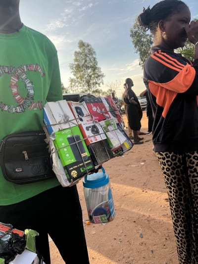 This image captures an outdoor street scene in Gbaza, Nigeria, under daylight conditions with clear skies and scattered clouds. In the foreground on the left, an individual is partially visible from the upper torso to the thighs. This person wears a bright green short-sleeved t-shirt adorned with a heart-shaped "CHANEL" logo composed of rectangular patches, and black trousers. A black fanny pack, bearing a "GG" logo resembling Gucci and "STAR" branding, is worn across their body. The individual holds a substantial bundle of packaged electronics, including power banks, earphones, and charging accessories (brands like "oraimo" and "Anifast" are visible, with one power bank listed as "15W TYPE-C FAST CHARGING," "20000mAh," and priced at "N 20000"). This bundle is secured with white straps. In their other hand, a clear plastic container with a blue lid, holding coiled cables labeled "Data CABLE," is visible.

To the right, in the mid-ground, a woman is partially visible from the chest downwards, with her head and shoulders in profile. She has dark skin, and her hair is styled in dark dreadlocks or braids pulled back into a bun. She wears a dark blue or black track jacket with distinct orange stripes on the sleeves and an orange lining, paired with leopard-print pants. Her right hand is raised near her face, possibly scratching or shading her eyes. A small, light-colored earring is visible on her ear.

The background features several indistinct figures, including one man in dark attire standing facing right. A light-colored vehicle is partially visible further back. The ground consists of reddish-brown, unpaved dirt with scattered pebbles and debris. A tall, slender tree with sparse foliage stands in the mid-ground, and other background vegetation is present. The lighting indicates direct sunlight, casting subtle shadows.