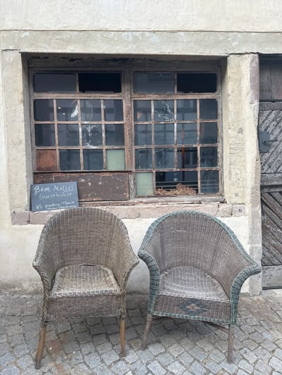 In Vianden, Luxembourg, two wicker chairs are placed in front of a weathered window with a rusted metal frame. A small chalkboard sign rests on the windowsill, displaying handwritten text that reads "BEIM MULLERS Guesthouse No parking Please maybe 0352-691392737". The building's facade is rough concrete, and the ground is paved with cobblestones. A wooden door with a diagonal plank pattern is visible to the right of the window. The scene appears to be an exterior view of a guesthouse.