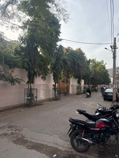 This image depicts a residential street scene under an overcast sky. On the left, a long, light-colored boundary wall runs parallel to the road. Along this wall, several tall, slender trees with dense green foliage are planted, each encased in a green metal protection cage at its base. A prominent wooden gate, arched at the top, is set into the wall, with a single brown chair positioned directly outside it. Above the wall, dense greenery, including plants with visible pink flowers, and the upper section of a building are present.

The asphalt road in the foreground and mid-ground shows signs of wear, with minor debris visible on its left edge. Two motorcycles are parked on the right side of the road; the closest one is black and red, identifiable as a Honda SP 125. Further down the street, a person riding a dark-colored scooter moves away from the viewer. A black sedan, bearing the license plate "DL 3CCP 1118" (indicating Delhi registration), is parked on the right side of the road. Two men are seen walking on the right, near the parked sedan. A utility pole supporting a street lamp and multiple overhead wires stands on the far right. In the distant background, low hills or mountains are faintly visible beneath the hazy sky.

The scene is characteristic of an urban or semi-urban street in India. While the prompt indicates the location as Jaipur, India, the visible license plate on the parked sedan specifies Delhi registration.