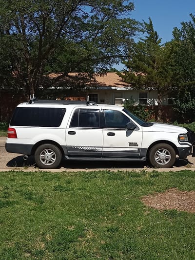 This media file is a full side-view, medium shot of a white, extended SUV parked on a residential street. The SUV is the primary subject, positioned in the center of the frame. It is a sunny day, and the sky is a clear blue. The vehicle appears to be a Ford Expedition, as indicated by the "Expedition" text visible on the side.
The SUV is white with a dark gray lower body trim that includes a decorative stripe graphic. It has tinted windows and a roof rack. The front of the vehicle has a black brush guard. The wheels are silver with a five-spoke design.
The SUV is parked on a concrete surface, likely a driveway or street, with a strip of green grass in the foreground. Behind the vehicle, a single-story house with a brown roof and white siding is visible, partially obscured by mature trees. The trees have full green foliage, suggesting it is summertime or late spring. The overall setting is a quiet, suburban neighborhood.
The image captures a static scene, with no people or obvious activity taking place. The lighting suggests it is mid-morning or early afternoon due to the bright sunlight and distinct shadows cast by the car. The location is identified as Amarillo, United States of America, which aligns with the appearance of the trees and architecture. The scene conveys a sense of normalcy and everyday life in a residential area.
