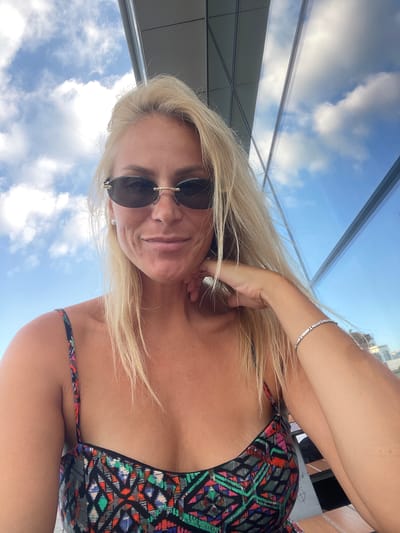 A woman with blonde hair and sunglasses is pictured in this media file. She is wearing a patterned bikini and a bracelet on her left arm. The scene appears to be outdoors, with a clear blue sky and clouds visible in the background, suggesting a daytime setting. The woman is looking towards the camera with a slight smile. The image is shot from a low angle, with the sky and a building or structure reflected in what appears to be a glass ceiling or window overhead. The overall mood is casual and relaxed, possibly indicating leisure time or a vacation. There is no visible text in the image.