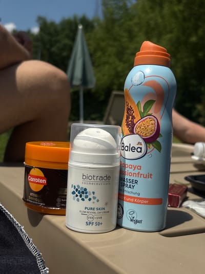 The photograph captures a close-up of several skincare products arranged on a surface, likely outdoors given the dappled sunlight and presence of greenery and a person's legs in the blurred background. The subjects in focus are three cosmetic items: an orange-lidded jar labeled "Carroten," a white pump bottle with "biotrade cosmeceuticals" visible, and a light blue aerosol can with "Balea" branding and an illustration of papaya and passionfruit. The environment suggests a sunny day, possibly in a recreational area, with a partial view of a green umbrella and trees. The image is composed from a slightly elevated angle, focusing on the products. The location is identified as Ivanyane, Bulgaria.