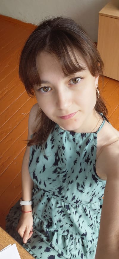 The image shows a young woman lying down on an orange wooden floor, looking up at the camera. She has long, brown hair with some lighter streaks, and is wearing a light teal dress with a black abstract pattern. She is also wearing a thin gold necklace and a gold hoop earring in her visible ear. On her left wrist, she has a white fitness tracker and a thin red string bracelet. Her expression is a slight, gentle smile. The background shows a light-colored wooden cabinet or desk and the textured orange floor. The lighting suggests it is daytime, possibly indoors. No text is visible in the image. The overall mood is casual and relaxed.