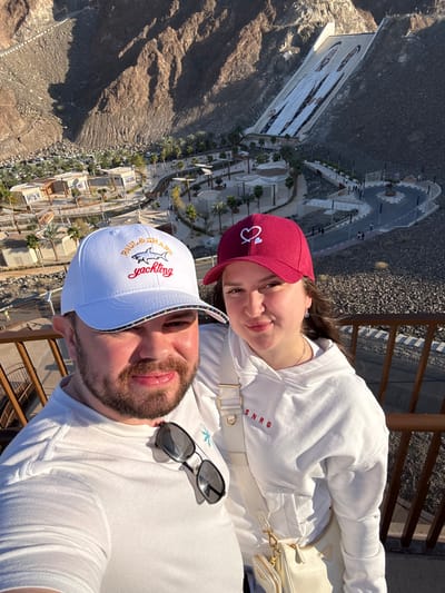The image depicts two individuals, a male and a female, positioned in the foreground from an elevated viewpoint in Hatta, United Arab Emirates, overlooking the Hatta Dam and its surrounding recreational area under clear daylight.
The male, on the left, is Caucasian with a beard and wears a white baseball cap featuring a 'Paul & Shark yachting' logo and a white t-shirt with sunglasses hooked onto the collar. He is smiling slightly towards the camera. The female, on the right, is Caucasian and wears a maroon baseball cap with a white heart and arrow emblem, a white hooded sweatshirt displaying 'SNRG' in red text, and carries a cream-colored shoulder bag with gold hardware. She is looking towards the camera with a subtle smile.
In the background, the prominent Hatta Dam is visible, characterized by a large concrete wall spanning a valley between rugged brown mountains. Two oversized, stylized white figures, resembling men in traditional attire, are painted vertically on the dam wall. Below the dam, a developed recreational zone features multiple low-rise buildings with light roofs, interconnected by paved pathways and a winding asphalt road. Palm trees and sparse vegetation are distributed throughout this area. Small, indistinct human figures are visible on the pathways and roads in the midground, indicating activity. The surrounding terrain consists of arid, rocky mountain slopes under direct sunlight, casting shadows. A brown metal railing of a viewing platform is partially visible in the immediate foreground.