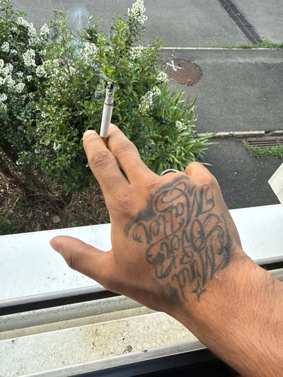 A person's hand, tattooed with script, holds a lit cigarette. The hand is positioned in the foreground, in front of a white windowsill. Behind the windowsill, a leafy green bush with small white flowers is visible. Beyond the bush, a grey paved area with a circular metal manhole cover and drainage grates can be seen, suggesting an urban outdoor setting. The lighting indicates it is daytime. The location is identified as Massy, France.