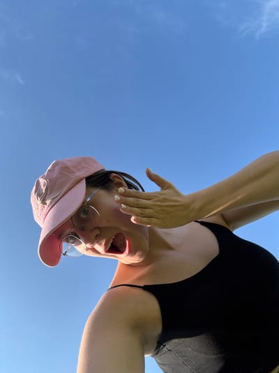 The image shows a woman with fair skin and dark hair wearing a pink baseball cap and a black tank top. She is wearing round glasses and has her mouth open, appearing surprised or yelling. Her right hand is raised, partially covering her face. The background is a clear blue sky with a few wispy clouds. The woman is positioned in the foreground, looking upwards at the camera, which is angled from below. The lighting suggests it is daytime. The image does not provide any information about a specific location in Dokshitsy, Russia.