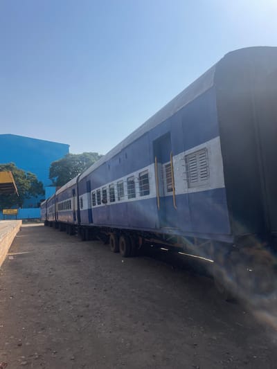 The image shows a blue and white train car at the Anajpur train station in Anajpur, India. The train is parked on a dirt surface, with a blue building and trees visible in the background.