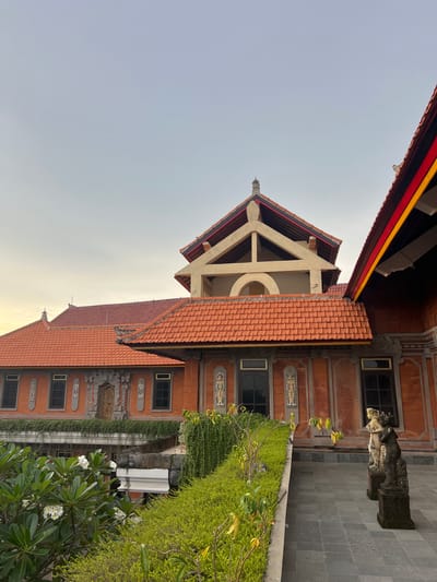 The image is a ground-level shot of Balinese architecture in Celuk, Indonesia. The frame is composed of a building with a red-tiled roof and orange walls, adorned with decorative carvings and rectangular windows. A separate roof structure, with a light-colored support beam, adds architectural complexity. Green shrubbery and a frangipani tree with white flowers grow along the base of the building. A stone statue sits on a paved stone surface. The sky is a pale gradient of light blue.