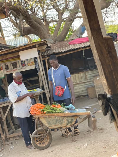 Two men are standing in a street market. One man is wearing a blue shirt and a red crossbody bag, standing near a wheelbarrow filled with carrots. The other man is wearing a white shirt and holding a piece of paper. They are positioned in front of a small wooden structure with a corrugated metal roof. Trees and various items are visible in the background. The photograph appears to be taken in a low-income or developing area.