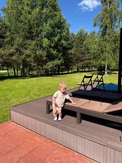This image captures an outdoor scene in Копнино, Russia, under bright sunlight. In the immediate foreground on the left, a section of red, square-patterned paving tiles is visible. Adjacent to this, occupying the lower half of the frame and extending to the right, is a raised platform constructed from dark brown composite or wooden decking material.
On this platform, a young child with curly blonde hair is positioned towards the right side of the frame, facing left. The child is wearing a light-colored t-shirt, a white diaper, and blue and white striped socks, and is standing upright with their right hand resting on a large, brown woven outdoor lounge chair equipped with brown cushions. Further back on the platform, two dark brown wooden folding chairs are present, alongside a blue tarp or cover.
The background consists of a vast, meticulously manicured green lawn. Beyond the lawn, a dense forest line features a mix of tall conifer trees and deciduous trees with vibrant green foliage. In the far left background, a hammock is suspended between two trees, with a person partially discernible resting within it. The sky is clear and blue, accented by a few scattered white cumulus clouds. Strong shadows are cast across the decking and lawn, indicating direct, intense sunlight.