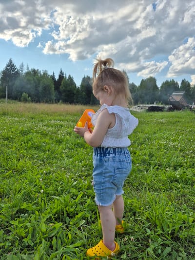 A young girl, viewed from the rear and right side, stands in a grassy field holding an orange toy gun. She is wearing a white ruffled top with pink polka dots, denim shorts, and yellow Crocs. Her hair is pulled into a high ponytail. The background features a line of trees, smoke rising from the left side, and what appears to be a rustic structure or debris pile on the right. The sky is partly cloudy. This scene is located in the village of Doyna, Russia.