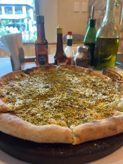 A high-angle, close-up shot features a pizza topped with what appears to be pistachio and shredded cheese. The pizza is placed on a dark-colored wooden board. Behind the pizza, various bottles are visible, including oil and sauce bottles. The scene is positioned indoors, near a window offering a glimpse of the outside environment, which seems to be a garden or street. The location in the city of Domingo Arenas, Mexico, is not directly identifiable from the image.